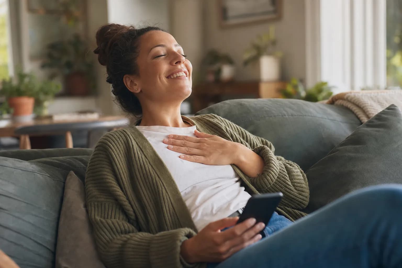 Femme soulagée, adossée à son sofa, téléphone à la main, dans un salon éclairé par la lumière du jour.